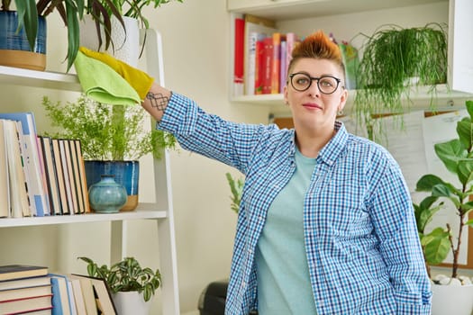 Middle-aged woman cleaning house, female looking at camera, wiping dust in room. Housekeeping, housework, housecleaning, cleaning concept