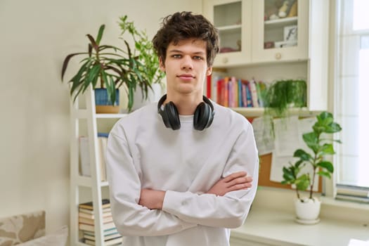Portrait of young handsome guy with crossed arms, in home interior. Smiling confident male 19-20 years old with curly hairstyle looking at camera. Lifestyle, youth concept