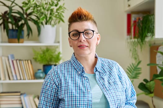 Portrait of smiling middle-aged woman in glasses with red haircut looking at camera in home interior. Mature people, lifestyle, health, life concept