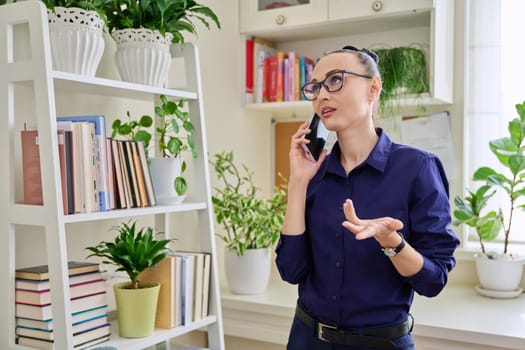 Beautiful emotional woman in her 30s talking on a cell phone, standing in her room at home. Conversation, communication, work, leisure, lifestyle, people concept