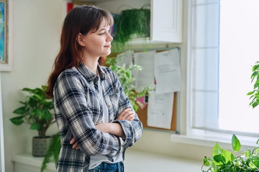 Smiling young woman looking out the window, in home interior, thinking dreaming worrying female, profile view. Lifestyle, feelings, mental health, lifestyle, youth concept