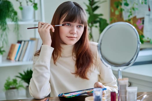 Young woman with cosmetic products on table, doing makeup looking in mirror at home. Cosmetics, youth, lifestyle, beauty concept