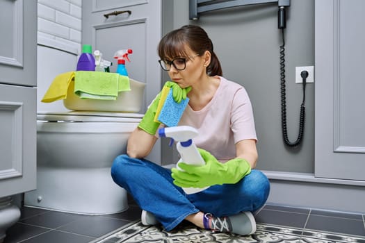 Tired woman sitting on the floor after cleaning bathroom, toilet. Routine house cleaning, home hygiene, housecleaning service, housekeeping, housework