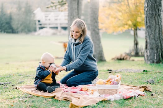 Mom gives a drink from a mug to a little boy sitting on a blanket in a clearing. High quality photo