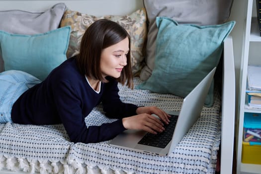 Young woman using laptop computer, typing on keyboard, lying on sofa at home. Internet online technologies for work communication study leisure