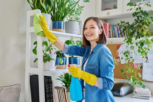 Young woman doing house cleaning, wiping dust, wearing gloves with cleaning detergent agent spray rag in room interior. Routine home cleaning, cleanliness housework housekeeping household lifestyle