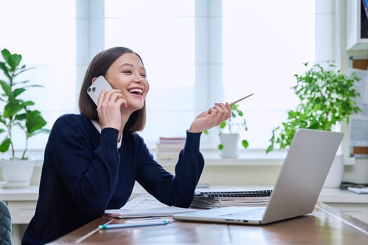 Happy smiling young woman sitting at desk at home office with computer laptop and talking on smartphone. Female working, studying remotely, freelancer, teacher, student