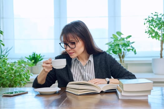 Middle-aged woman reading books sitting at home at a table with cup of coffee. Literary hobby, leisure, relaxation, people concept