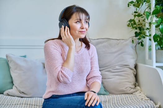 Middle-aged woman in headphones listening to audio, music, audiobook, communication, relaxed vacationer sitting on couch at home