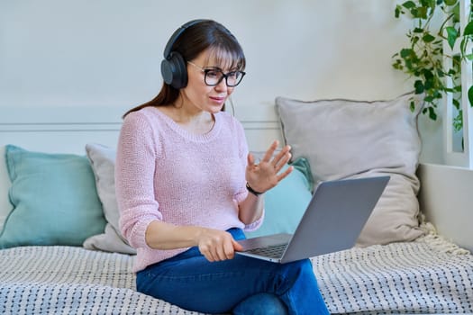 Middle aged woman in headphones having video call chat conference on laptop sitting on couch at home. Vlog, work, leisure, online communication, modern technologies concept