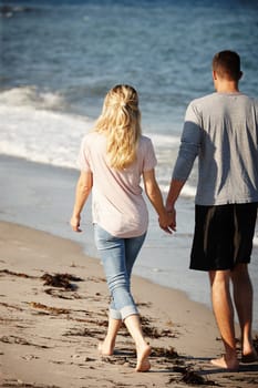 Couple, walking and holding hands on shore of beach, peace and travel to nature for holiday. People, back and calm stroll on weekend or vacation, love and date for relationship or marriage and ocean.