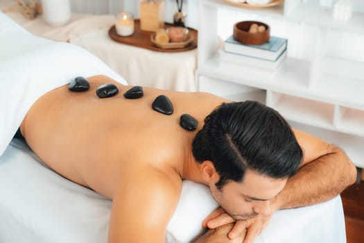 Hot stone massage at spa salon in luxury resort with day light serenity ambient, blissful man customer enjoying spa basalt stone massage glide over body with soothing warmth. Quiescent