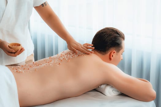 Blissful man customer having exfoliation treatment in luxury spa salon with warmth candle light ambient. Salt scrub beauty treatment in health spa body scrub. Quiescent