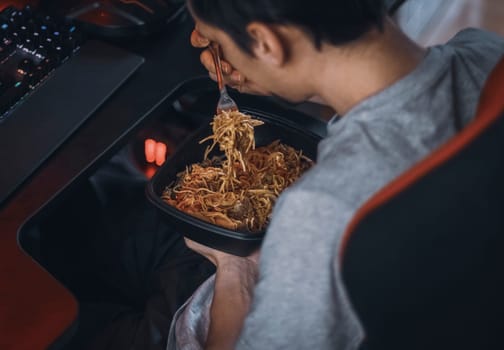 Young caucasian guy gamer eats wok food with a fork from a black container during lunch break,sitting in a work chair at a table with a keyboard,top view close-up with depth of field.Concept of food process,wok,fast food,using technology,dark style,office lunch .