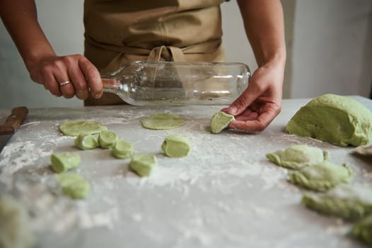 Close-up housewife, woman chef in beige apron, using an empty glass wine bottle as rolling pin, rolling out dough while cooking dumplings at rustic home kitchen. Female chef preparing green ravioli