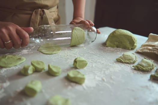 Close-up hands of woman in beige apron, using a glass wine bottle for rolling out the dough with green spinach, cooking traditional Ukrainian Varennyky with mashed potatoes. Food. Traditions. Culture