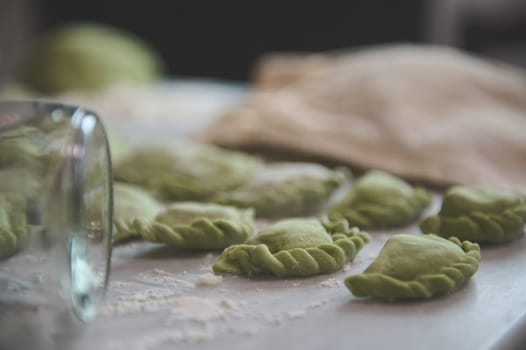 Close-up homemade dumplings of green color on a floured kitchen table and partial view of a glass bottle as rolling pin on the foreground. Preparing traditional cuisine in Ukraine - varennyky