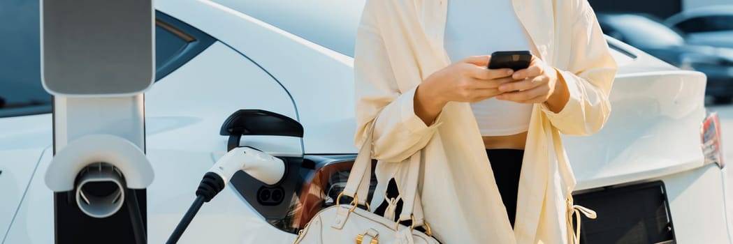 Woman holding shopping bag and use smartphone to pay for electricity to recharge EV car battery from charging station at city mall parking lot. Modern woman go shopping by eco car.Panorama Expedient