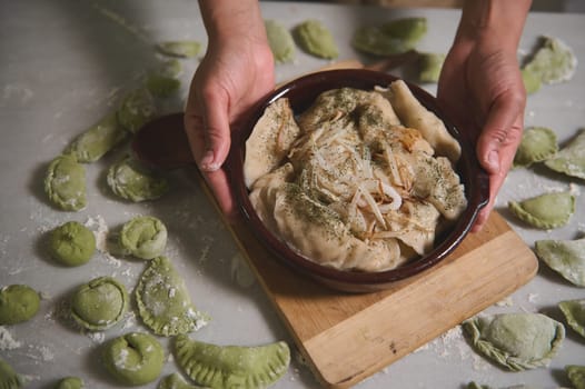 Close-up of a housewife, chef putting a clay dish with freshly boiled Ukrainian varennyky with roasted onions on a floured table with molded dumplings. Traditional Ukrainian cuisine. Homemade culinary