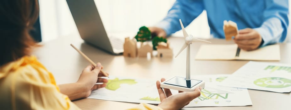 Cropped image of businesswoman presents eco-friendly house by using green design to manager while holding the wooden house block at table with windmill model placed with document. Delineation.