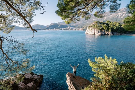 View through the green branches of a man on a stone ledge above the sea, raising his hands up and looking at the mountains. Back view. High quality photo