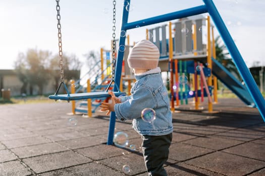 Little girl stands near a swing and catches soap bubbles. Side view. High quality photo