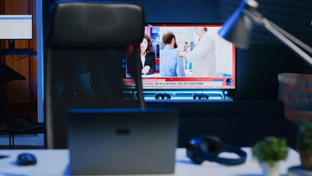 Smart TV left open on news broadcast in empty home office with desk filled with business items. Television set showing VOD channel with Laptop, headphones, plants in blurry foreground, camera A