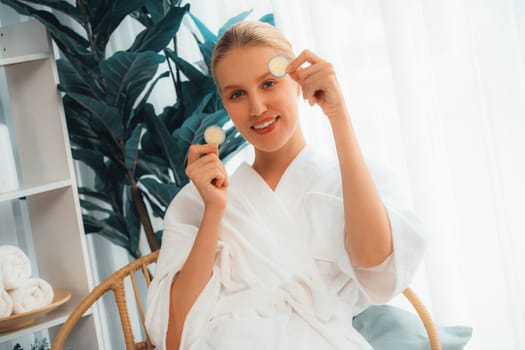 Woman holding slices of fresh cucumber and wearing bathrobe enjoying luxurious facial skincare spa in resort or hotel. Skin treatment for face and beauty care. Quiescent