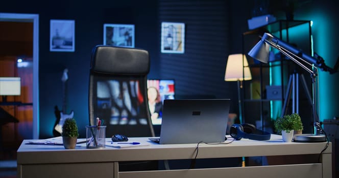 Focus on office desk in front of smart TV in blurry background displaying news program broadcasting. Laptop on table and television screen in empty room showing VOD channel, camera B