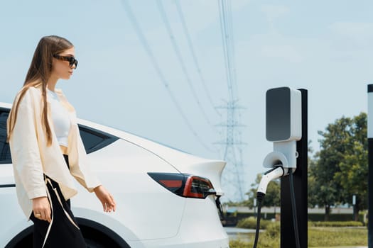 Young woman recharge EV car battery at charging station connected to power grid tower electrical industrial facility as electrical industry for eco friendly vehicle utilization. Expedient