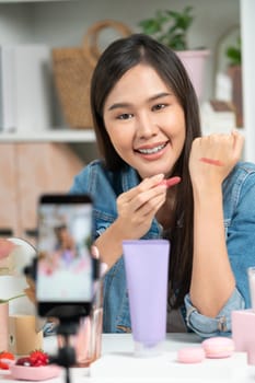 Young beautiful Asian showing cosmetic makeup lipstick matte pastel color testing in bestseller collection on arm test promotion on social media online recording smartphone at modern room. Stratagem.