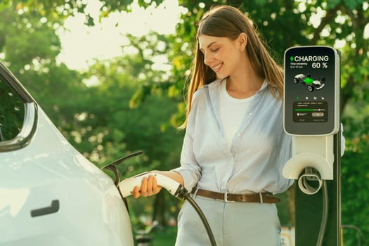 Young woman recharging battery for electric car during road trip travel EV car in natural forest or national park with sunrise lighting. Eco friendly travel during vacation and holiday. Exalt