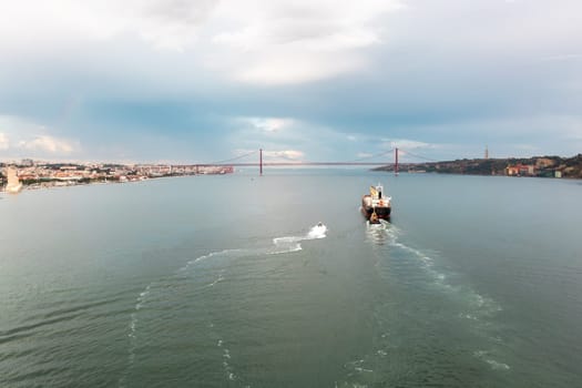 Aerial drone shot of cargo ship moving in Tagus river with Ponte 25 de Abril in background. The iconic Ponte 25 de Abril spans the background, set against the backdrop of a cloudy sky.