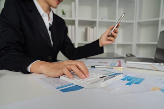 A woman in a business suit is using a calculator and a cell phone. She is focused on her work and she is in a professional setting