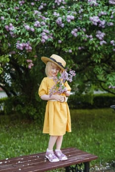 A little girl in a yellow dress and straw hat wearing a bouquet of lilacs. A walk in a spring park, blossoming lilacs