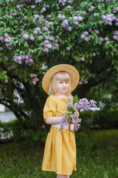 A little girl in a yellow dress and straw hat wearing a bouquet of lilacs. A walk in a spring park, blossoming lilacs