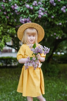 A little girl in a yellow dress and straw hat wearing a bouquet of lilacs. A walk in a spring park, blossoming lilacs