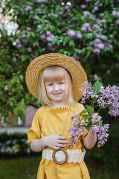 A little girl in a yellow dress and straw hat wearing a bouquet of lilacs. A walk in a spring park, blossoming lilacs