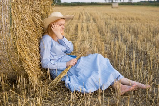 A red-haired woman in a hat and a blue dress walks in a field with haystacks