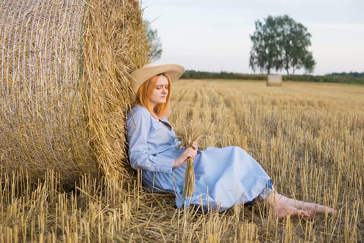 A red-haired woman in a hat and a blue dress walks in a field with haystacks