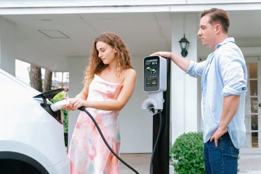 Happy and lovely couple with eco-friendly conscious recharging electric vehicle from EV home charging station. EV car technology utilized for residential home to future sustainability. Synchronos