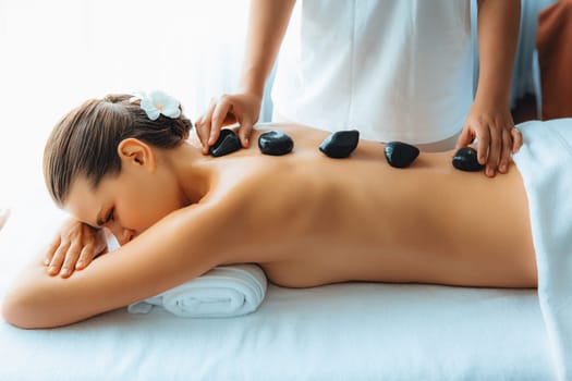 Hot stone massage at spa salon in luxury resort with day light serenity ambient, blissful woman customer enjoying spa basalt stone massage glide over body with soothing warmth. Quiescent