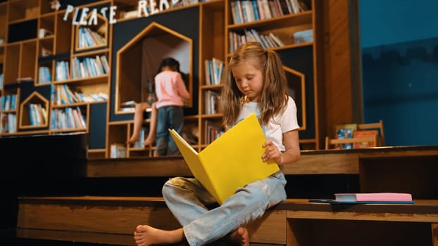 Young smart caucasian girl picking reading a book while sitting at library. Clever child learning, studying, open a books at library. Attractive kid turning page with blurring background. Erudition.