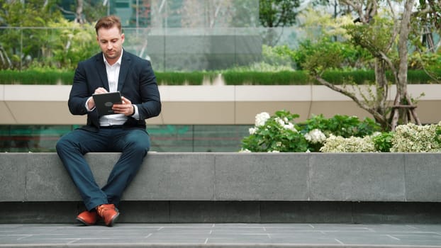 Caucasian smart businessman sitting at park while writing on tablet by using tablet pencil and planning marketing strategy. Manager looking at data analysis while thinking about financial plan. Urbane