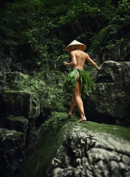 A naked sexy girl in a triangular Asian hat and a homemade cape made of leaves walks along the canyon of mountain river