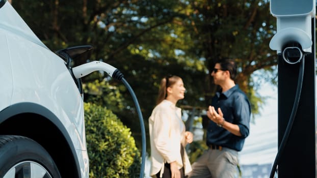 Young couple recharge electric car's battery from charging station in outdoor green city park in springtime. Rechargeable EV car for sustainable environmental friendly urban travel lifestyle.Expedient