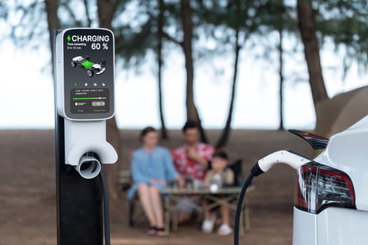 Alternative family camping trip traveling by the beach with electric car recharging battery from EV charging station with blurred family enjoying the seascape campsite background. Perpetual