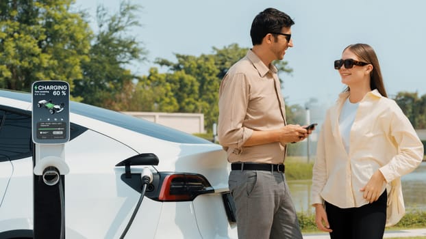 Young couple use smartphone to pay for electricity at public EV car charging station green city park. Modern environmental and sustainable urban lifestyle with EV vehicle. Expedient