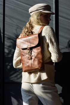 Studio portrait of beautiful woman with a curly blond hair holding brown backpack, posing on gray background. Model wearing stylish cap, sweater and classic trousers