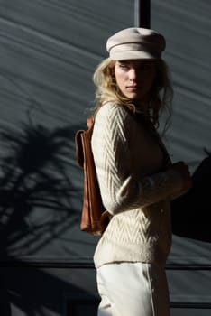 Studio portrait of beautiful woman with a curly blond hair holding brown backpack, posing on gray background. Model wearing stylish cap, sweater and classic trousers